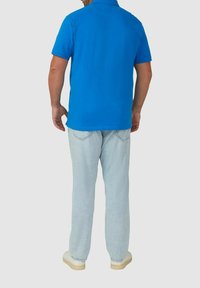Mężczyzna w jasnoniebieskiej koszulce polo z krótkim rękawem, jasnoniebieskich dżinsach i białych trampkach, stoi plecami do prostego tła.
