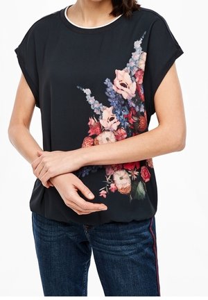 Kvinde iført sort top med korte ærmer med pink, rød og blå blomsterprint på højre side, kombineret med blå jeans med sidestribe.