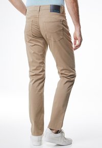 Beige slim-fit housut, joissa on sileä pinta, viisi taskua ja musta merkki vyötäröllä, yhdistettyinä valkoisiin lenkkareihin.