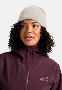 Vrouw met krullend haar, die een beige fleece muts en een geplooide pruimen Jack Wolfskin jas draagt, en iets opzij kijkt.