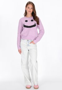 Paarse fluffy trui met een zwart smiley-ontwerp. Gecombineerd met lichtblauwe wijde jeans en roze schoeisel.