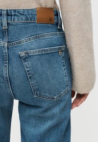 Κοντινό πλάνο ατόμου που φοράει μπλε τζιν τζιν με ταμπά δερμάτινο έμπλαστρο και μπεζ πουλόβερ, εστιάζοντας στην πίσω τσέπη και το χέρι στο πλάι.