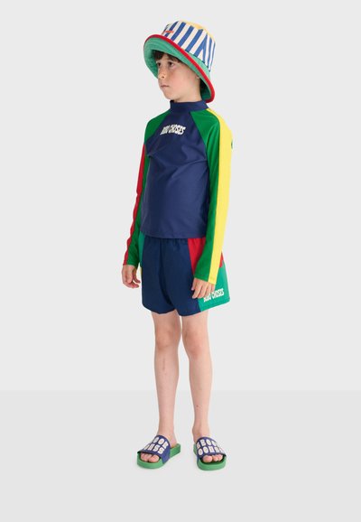 Child standing, wearing a colorful long-sleeve rash guard, matching shorts, striped bucket hat, and blue slide sandals with text.