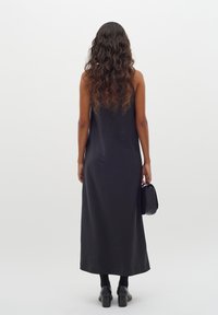Czarna bezrękawnikowa sukienka maxi o gładkiej fakturze, w połączeniu z małą czarną torebką. Modelka nosi czarne botki do kostek i rajstopy.