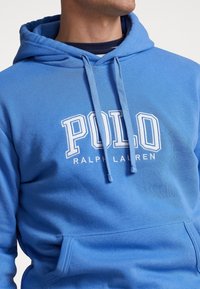 Man som bär en blå Polo Ralph Lauren-hoodie med dragsko och kenguruficka, avskuren vid halsen och midjan.
