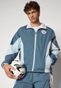 Giacca a vento blu chiaro e blu scuro con dettagli bianchi, chiusura con zip, logo sportivo, indossata da una persona che tiene un pallone da calcio bianco e nero.