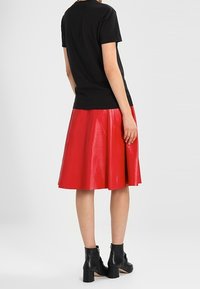 Person wearing a black short-sleeve top, red leather knee-length skirt, and black ankle boots facing away on a plain background.