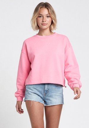 Tânără cu păr scurt și blond, purtând un hanorac roz larg și pantaloni scurți din denim albastru deschis, stând în fața unui fundal simplu.