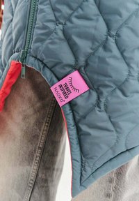 Prešívaná bunda s tyrkysovým vonkajškom a červeným vnútrom. Má ružovú nášivku s nápisom „TRAVEL INSPIRED“ pri zipsu.