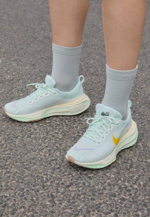 Person trägt hellblaue Nike-Laufschuhe mit gelben und lavendelfarbenen Swoosh-Logos auf grauem Asphaltboden.