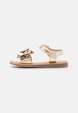 Metallisch goldene Sandalen mit flacher Sohle, ausgestattet mit einer blumigen Verzierung am Riemen und einem verstellbaren Knöchelverschluss.