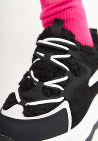 Baskets texturées noir et blanc avec des lacets bouclés uniques, portées avec des chaussettes rose vif sur un fond clair.