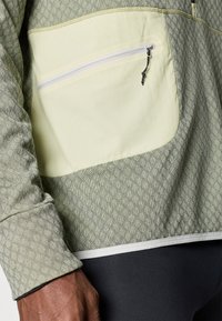 Szürke és világoszöld textúrált pulóverdzseki cipzáros zsebbel és kontrasztos díszítéssel. Mintás anyag a további részletekért.