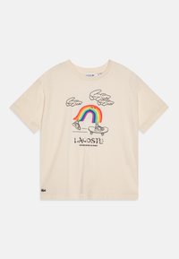 Krémové bavlněné tričko s hravým grafickým designem, který zobrazuje duha, mraky a tenisky. Obsahuje text a logo "Lacoste".