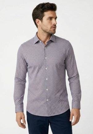 Mann trägt ein schlank geschnittenes, langärmliges Hemd mit kleinem Karomuster und dunkle Hose, schaut zur Seite.