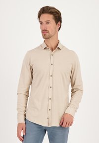 Camisa de manga larga beige con cierre frontal de botones y botones color oliva. Cuenta con un cuello clásico y un logotipo sutil en el pecho. Textura suave.