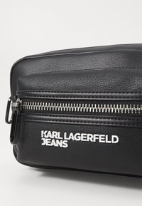 Schwarze Ledertasche mit strukturiertem Oberflächen, ausgestattet mit einem silbernen Reißverschluss und einem weißen Markenlogo. Kompakte rechteckige Form, dezente Akzente.