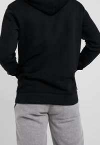 Felpa con cappuccio nera dalla texture liscia, con tasca a marsupio e polsini a coste. Abbinata a pantaloni della tuta grigi in tessuto morbido.