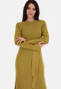 Abito in maglia a costine verde oliva con scollo rotondo e maniche lunghe, decorato con dettagli a fiocco sui polsini. Silhouette semplice e aderente.
