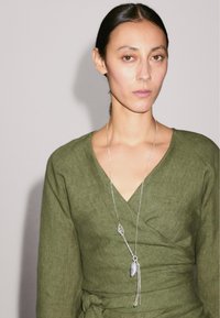 Une jeune femme aux longs cheveux noirs porte un haut vert à enrouler et un collier en argent avec un pendentif, se tenant devant un fond clair.