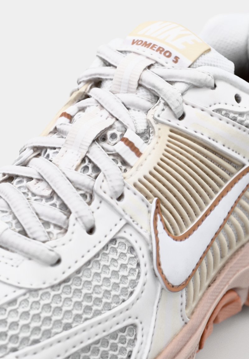 Nike Vomero 5 sneaker in wit en beige, met een mesh bovenwerk, ronde neus en textuuraccenten. Witte veters en een kenmerkend swoosh-logo.