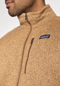 Brūna vilnas jaka ar augstu apkakli, priekšējo rāvējslēdzēju un Patagonia logotipu. Materiāls ir ar teksturētu, jauktu toņu virsmu.