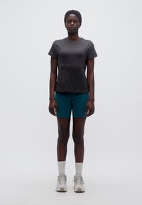 Άτομο που στέκεται με κοντά μαλλιά φοράει εφαρμοστό μαύρο t-shirt, γαλάζια ποδηλατικές βερμούδες, λευκές κάλτσες μέχρι τον αστράγαλο και γκρίζα αθλητικά παπούτσια μπροστά σε απλό φόντο.