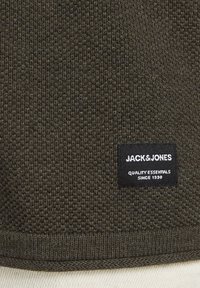 Tissu en maille texturé vert olive foncé avec une étiquette noire rectangulaire portant la mention "JACK & JONES Quality Essentials Since 1990".