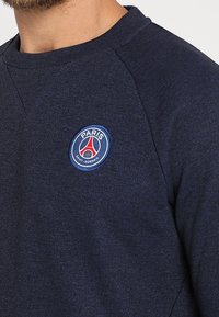 Sweat-shirt bleu marine avec manches raglan, arborant un logo rond brodé en bleu et rouge pour le Paris Saint-Germain sur la poitrine.