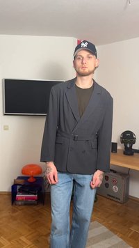 Ciemnoszara marynarka dwurzędowa z paskiem w talii, kołnierzem z klapami i smukłym krojem, noszona na oliwkowej zielonej koszuli i jasnych dżinsach.