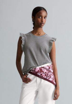 Femme portant un haut rayé sans manches avec des volants aux épaules, un pantalon blanc et une écharpe rose à motifs nouée autour de la taille.