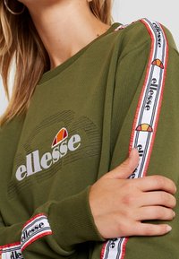 Sudadera verde oliva con puños y cuello de canalé, que presenta un logo bordado en blanco y una cinta lateral a rayas rojas y blancas con marca.