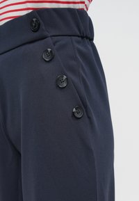 Granatowe spodnie z teksturowanej tkaniny, z wysokim stanem, bocznymi guzikami i zwężanym krojem. Przód zdobią czarne guziki.