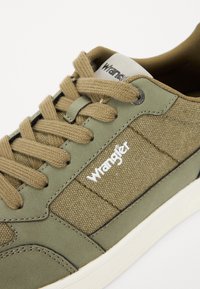 Sneakers verde oliva con tomaia in tessuto testurizzato, lacci color beige e logo "Wrangler" bianco. Presentano un design elegante e una suola in gomma.