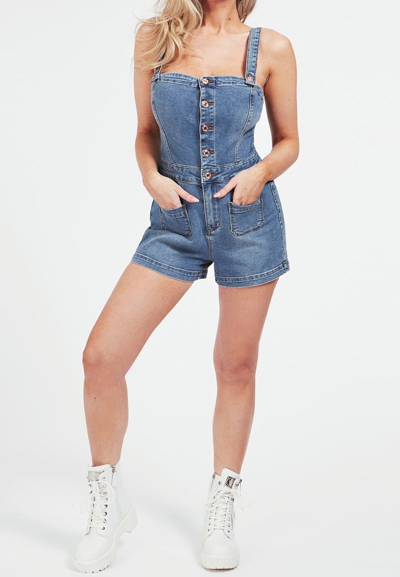 Femme portant une combinaison courte en denim bleu boutonnée avec poches avant et bottes blanches à lacets, debout les mains dans les poches sur fond blanc.