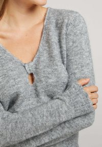 Pull en tricot gris avec un décolleté en V profond et un détail en trou de serrure. La texture est douce avec des poignets côtelés, mettant en avant une coupe décontractée.