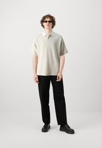 Ανοιχτό γκρι πόλο μπλουζάκι με ραβδωτή υφή και αντίθετο λευκό γιακά, συνδυασμένο με μαύρα παντελόνια σε φαρδιά γραμμή και μαύρα παπούτσια.