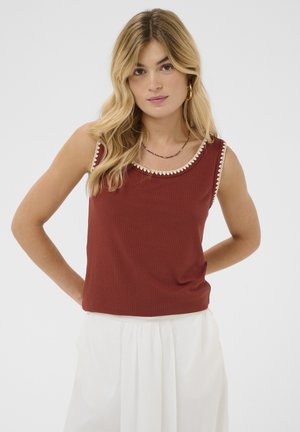 Jeune femme aux longs cheveux blonds portant un haut sans manches rouge rouille à bordure blanche et un pantalon taille haute blanc, posant devant un fond uni.