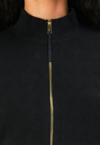 Svart zip-up jakke med høy krage, teksturert stoff og gullglidelås med et distinkt mønster av subtile blomsterdesign.