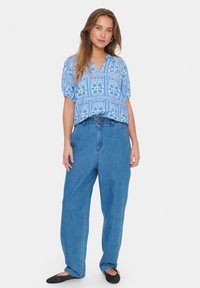Top blu con motivo, scollatura a V e maniche corte, abbinato a pantaloni in denim blu larghi e scarpe nere basse.