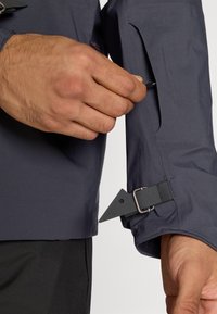 Vandeniui atsparus pilkas striukės rankovės bruožas - užtrauktukais užsegama kišenė, reguliuojama juosta su metaliniu užsegimu ir struktūruota medžiaga patvarumui.