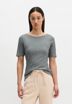 Kobieta o długich ciemnych włosach, ubrana w dopasowany szary top z krótkim rękawem i beżowe spodnie ze sznurkiem, stojąca na jednolitym białym tle.