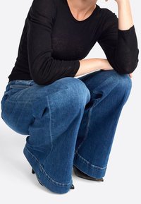 Weite blaue Denim-Jeans mit fransigem Saum, hoher Taille und zwei großen Seitentaschen. Kombiniert mit einem eng anliegenden schwarzen, gerippten Langarmoberteil.
