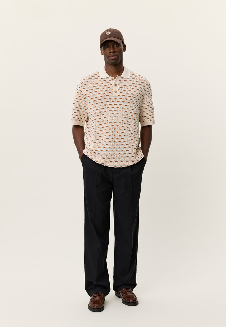Polohemd mit Muster aus farbigen Formen, beige Hintergrund; kombiniert mit schwarzen Hosen und braunen, strukturierten Schuhen; weißer Kragen. Braune Mütze.