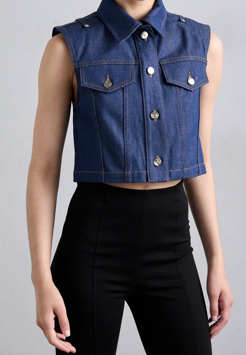 Veste courte en denim bleu sans manches avec fermetures à boutons, poches poitrine à rabat, portée avec un pantalon noir taille haute sur une personne debout.