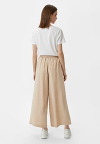 Weißes Baumwoll-T-Shirt und beige weite Hose mit elastischem Bund, kombiniert mit weißen Sneakers, von hinten betrachtet.