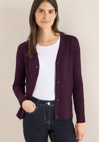 Bordeauxroter, gerippter Cardigan mit langen Ärmeln, fünf Knöpfen und einem leicht geschwungenen Saum. Trägt man über einem weißen T-Shirt und kombiniert mit dunklen Jeans.