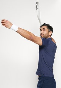 Polo desportivo azul-marinho com mangas curtas, tecido texturizado, usado com uma pulseira branca. A figura está a servir com uma raquete de ténis.