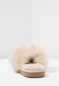 Pantoufle à glissière en fausse fourrure beige avec un dessus moelleux et duveteux, une semelle intérieure douce et une semelle texturée en tan. Bout carré et bords arrondis et duveteux.