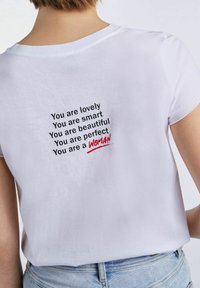 Camiseta de algodón blanca con cuello redondo que presenta texto negro en la parte posterior, afirmaciones con la palabra "MUJER" en rojo.